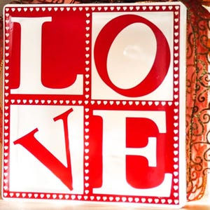 May include: A white ceramic square sign with a red "LOVE" written in block letters. The letters are outlined with a white dotted line of small hearts.