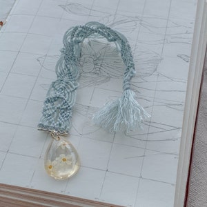 May include: A light blue macrame bookmark with a tassel and a clear teardrop-shaped charm containing pressed white flowers.