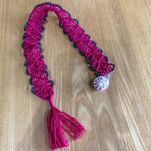 May include: A pink and black macrame bookmark with a shell charm and a tassel.