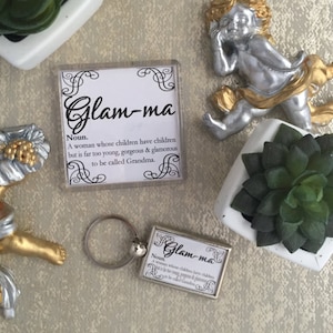 May include: A white square keychain with a black and white design that reads "Glam-ma" and defines the word as "A woman whose children have children but is far too young, gorgeous & glamorous to be called Grandma."