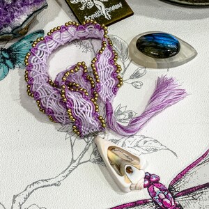 May include: A purple and white macrame bracelet with gold beads and a shell pendant. The bracelet has a tassel and is tied with a knot.