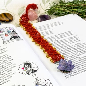 May include: Red and gold macrame bookmark with a purple butterfly charm. The bookmark is lying on a book with text about a witch's spell.