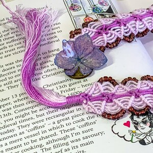 May include: A purple and white macrame bracelet with brown beads. The bracelet is decorated with a pressed flower.