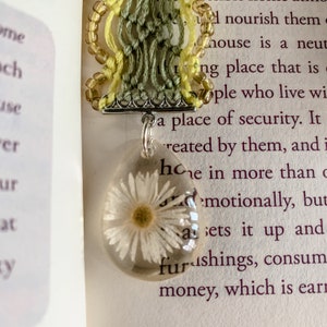 May include: A white daisy pressed inside a clear resin pendant, hanging from a macrame cord with green and yellow threads. The pendant is attached to a silver chain.