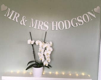May include: White banner with the text "MR & MRS HODGSON" and two small white hearts on either end. The banner is hanging on a light green wall. A white orchid plant is in a white pot on a white shelf with a string of white lights.
