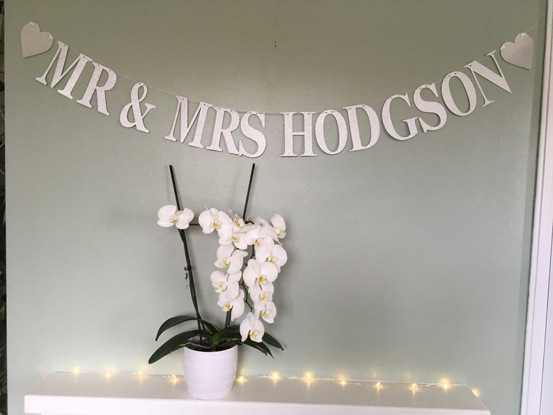 Puede incluir: Pancarta blanca con el texto "MR & MRS HODGSON" y dos peque&ntilde;os corazones blancos en cada extremo. La pancarta est&aacute; colgada en una pared verde claro. Una planta de orqu&iacute;dea blanca est&aacute; en una maceta blanca sobre una repisa blanca con una cadena de luces blancas.