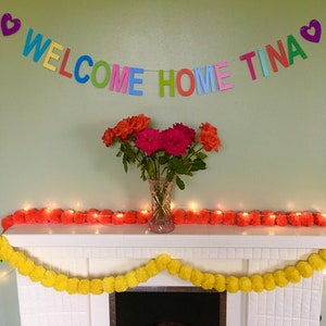 May include: A white fireplace mantel decorated with a colorful welcome home banner that reads "WELCOME HOME TINA" and two garlands of pom poms, one yellow and one orange. The banner is strung with hearts at each end. A vase of pink and orange roses sits on the mantel.