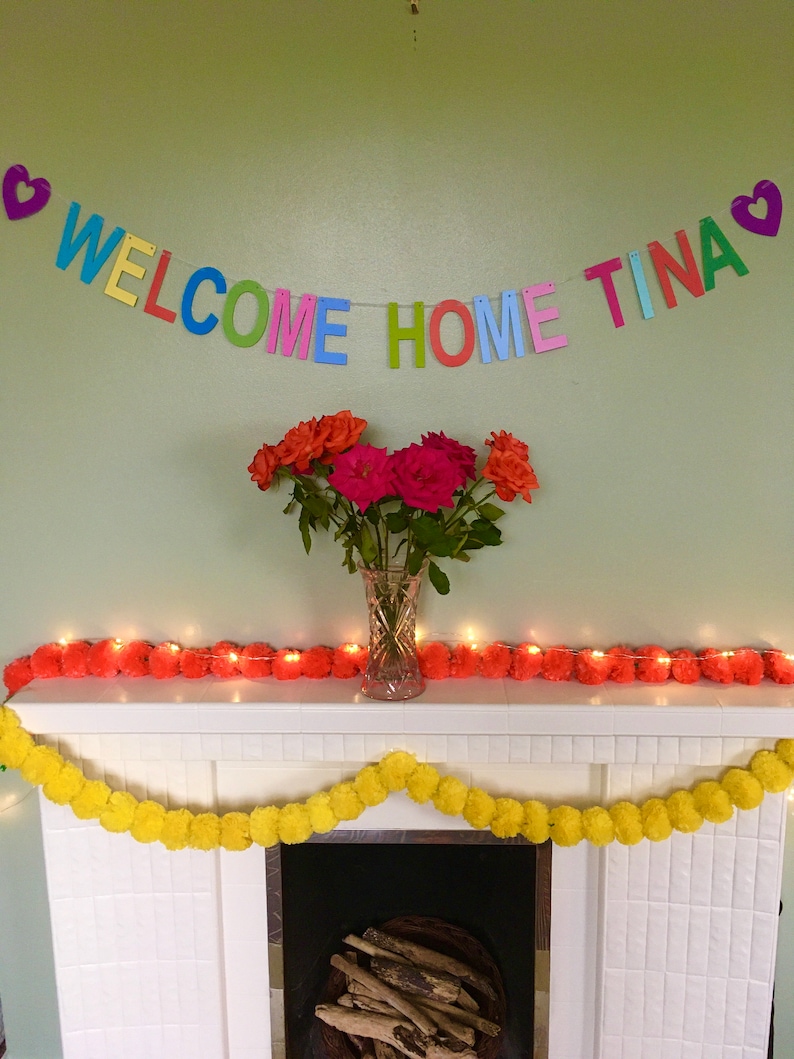 May include: A white fireplace mantel decorated with a colorful welcome home banner that reads "WELCOME HOME TINA" and two garlands of pom poms, one yellow and one orange. The banner is strung with hearts at each end. A vase of pink and orange roses sits on the mantel.