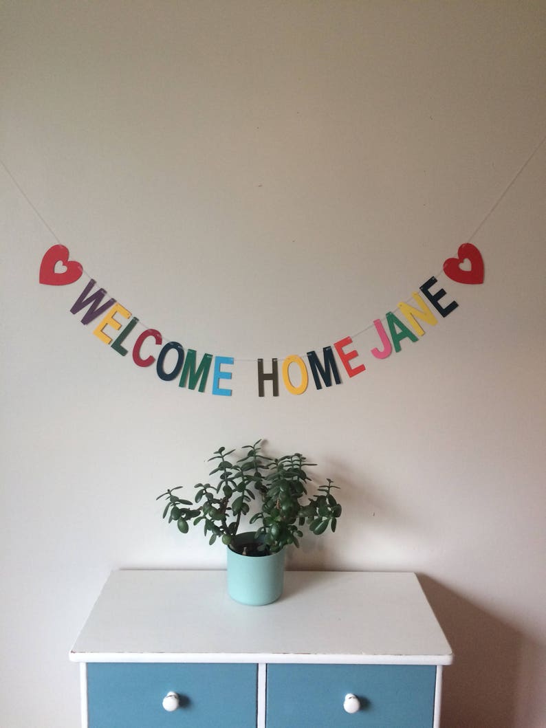 May include: A colorful welcome home banner with red hearts on each end and the words "WELCOME HOME JANE" in rainbow colors.