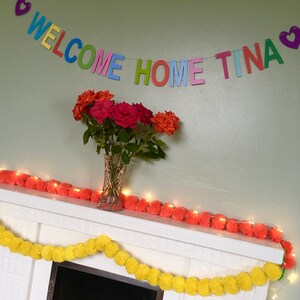 May include: A colorful welcome home banner with the words "WELCOME HOME TINA" and hearts on either end. The banner is hanging above a white fireplace mantel decorated with a garland of yellow and orange pom-poms.