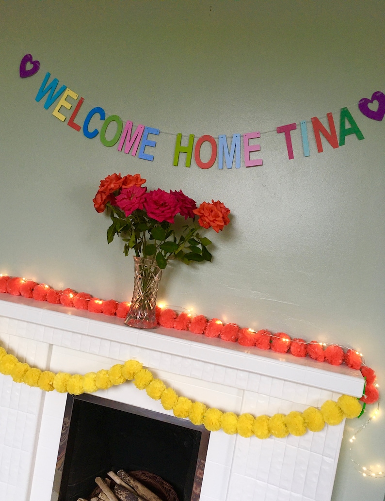 May include: A colorful welcome home banner with the words "WELCOME HOME TINA" and hearts on either end. The banner is hanging above a white fireplace mantel decorated with a garland of yellow and orange pom-poms.