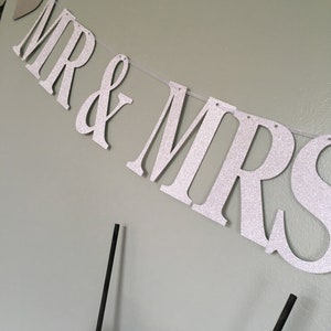 Puede incluir: Una pancarta de purpurina plateada con las palabras "MR & MRS" colgada en una pared. La pancarta est&aacute; enhebrada con hilo blanco y tiene un peque&ntilde;o coraz&oacute;n plateado al principio.