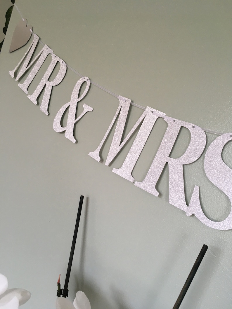 Puede incluir: Una pancarta de purpurina plateada con las palabras "MR & MRS" colgada en una pared. La pancarta est&aacute; enhebrada con hilo blanco y tiene un peque&ntilde;o coraz&oacute;n plateado al principio.