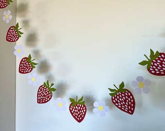 May include: A red and green paper garland with strawberries and white flowers. The garland is strung together with white string.