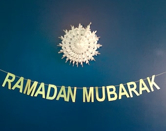 May include: A gold banner with the text "RAMADAN MUBARAK" hanging on a blue wall. A white paper snowflake is hanging above the banner.