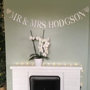 Puede incluir: Pancarta blanca con el texto "MR & MRS HODGSON" en letras blancas con corazones en cada extremo. La pancarta est&aacute; colgada sobre una chimenea blanca con un hogar negro. Una planta de orqu&iacute;dea blanca est&aacute; sentada en la repisa de la chimenea.
