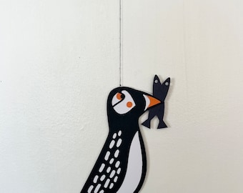 May include: A black and white paper cut-out of a puffin holding two fish in its beak. The puffin has orange feet and a white belly. The fish are black and white.