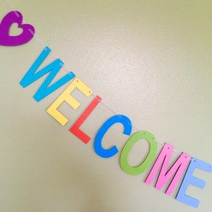 May include: A colorful welcome banner with a purple heart at the beginning of the string. The letters are made of felt and are in the colors blue, yellow, red, green, pink, and light blue. The letters spell out the word "WELCOME".