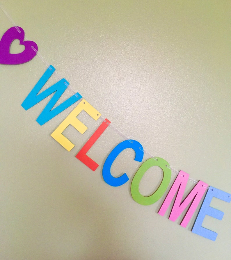 May include: A colorful welcome banner with a purple heart at the beginning of the string. The letters are made of felt and are in the colors blue, yellow, red, green, pink, and light blue. The letters spell out the word "WELCOME".