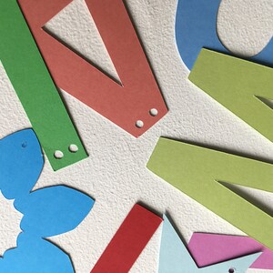 May include: Assortment of colorful paper cutouts, including letters, a flower, and geometric shapes. The cutouts have holes punched in them, suggesting they are part of a banner or garland.