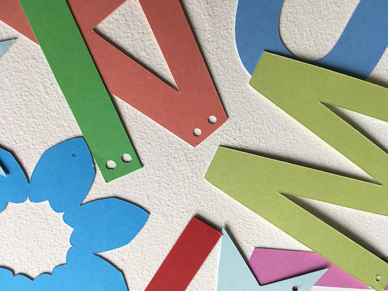 May include: Assortment of colorful paper cutouts, including letters, a flower, and geometric shapes. The cutouts have holes punched in them, suggesting they are part of a banner or garland.