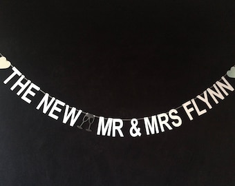 May include: White banner with the text "THE NEW MR & MRS FLYNN" with two small hearts and a pair of champagne glasses.