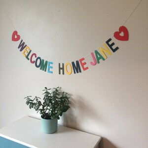 May include: A colorful welcome home banner with the words "Welcome Home Jane" and two red hearts. The banner is strung on a white string and is hanging on a white wall.