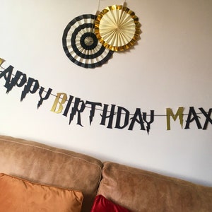 May include: Black and gold "Happy Birthday Max" banner with lightning bolt accents. The banner is hanging on a white wall with two paper fans above it.