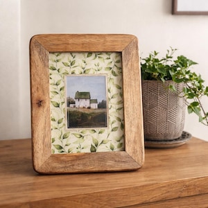 May include: A wooden picture frame with a rectangular shape and rounded corners. The frame displays a picture of a white house with a green roof. The picture is set against a green and white leaf pattern. The frame rests on a wooden surface.