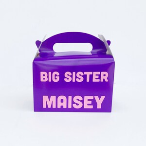 K&ouml;nnte beinhalten: Lilafarbener Geschenkbox mit Griff. Die Box hat den Text "BIG SISTER MAISY" in rosa Buchstaben auf der Vorderseite.