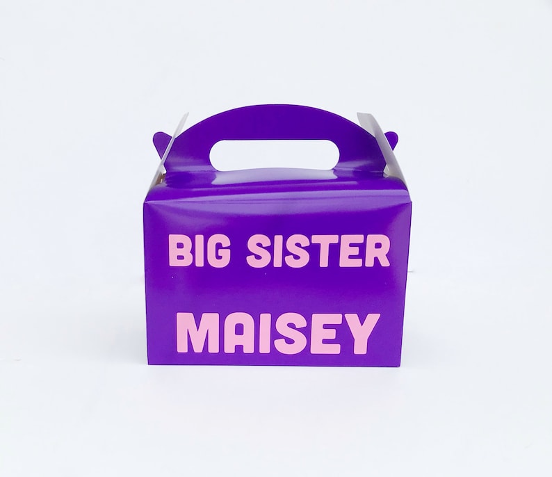 K&ouml;nnte beinhalten: Lilafarbener Geschenkbox mit Griff. Die Box hat den Text "BIG SISTER MAISY" in rosa Buchstaben auf der Vorderseite.