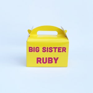May include: Yellow cardboard gift box with a handle. The box has the text "BIG SISTER RUBY" printed in pink.