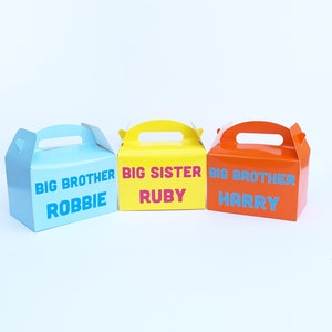 K&ouml;nnte beinhalten: Drei kleine Geschenkboxen mit Griffen. Die Boxen sind blau, gelb und orange. Die Boxen sind mit "Gro&szlig;er Bruder Robbie", "Gro&szlig;e Schwester Ruby" und "Gro&szlig;er Bruder Harry" beschriftet.