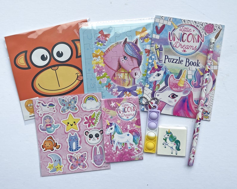 May include: A unicorn-themed activity pack for kids includes a puzzle book, stickers, a pencil, a pop-it toy, and a puzzle. The puzzle book cover features a unicorn with a rainbow mane and the words "Little Unicorn Dreams Puzzle Book".