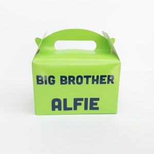 K&ouml;nnte beinhalten: Eine gr&uuml;ne Geschenkschachtel aus Pappe mit einem Griff. Auf der Vorderseite der Schachtel steht "BIG BROTHER ALFIE" gedruckt.