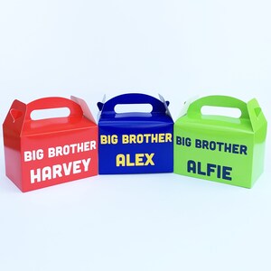 K&ouml;nnte beinhalten: Drei Geschenkboxen mit Griffen, jede in einer anderen Farbe: Rot, Blau und Gr&uuml;n. Die Boxen sind mit dem Text "Gro&szlig;er Bruder" und einem Namen verziert: Harvey, Alex und Alfie.