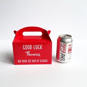 May include: A red cardboard gift box with a handle, personalized with the text "GOOD LUCK Thomas ON YOUR 1ST DAY AT SCHOOL". A silver can of Diet Coke is next to the box.