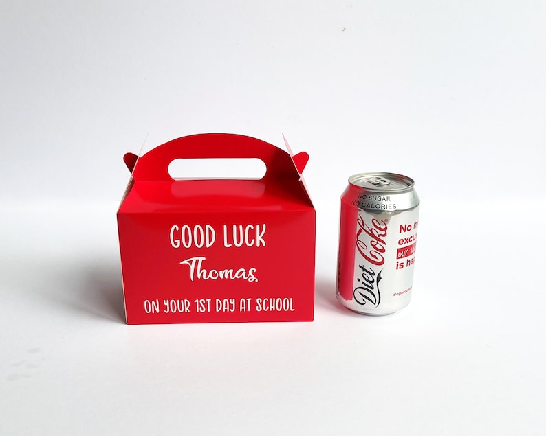 May include: A red cardboard gift box with a handle, personalized with the text "GOOD LUCK Thomas ON YOUR 1ST DAY AT SCHOOL". A silver can of Diet Coke is next to the box.