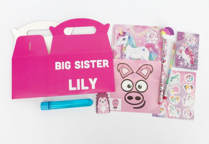 May include: Pink and white gift box with the text "Big Sister Lily" on the front. The box is filled with a variety of items, including a unicorn puzzle, a unicorn pencil, a unicorn sticker sheet, a pig face mask, a hedgehog eraser, and a blue bubble wand.