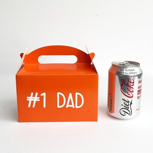 K&ouml;nnte beinhalten: Orangefarbener Geschenkkarton aus Pappe mit einem Griff und dem aufgedruckten Text "#1 DAD" auf der Vorderseite. Die Schachtel steht neben einer silbernen Dose Diet Coke.