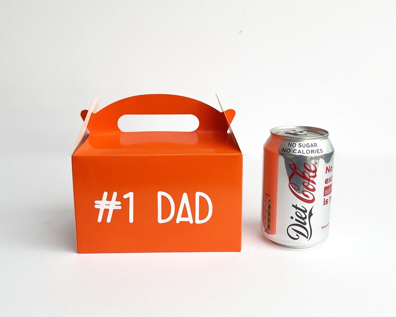 K&ouml;nnte beinhalten: Orangefarbener Geschenkkarton aus Pappe mit einem Griff und dem aufgedruckten Text "#1 DAD" auf der Vorderseite. Die Schachtel steht neben einer silbernen Dose Diet Coke.