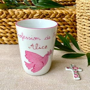 Puede incluir: Una taza de cerámica blanca con un borde rosa y una paloma rosa con una rama de olivo. La taza tiene el texto "Profession de Foi Alice" escrito en ella.