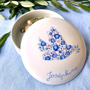 May include: White ceramic jewelry box with a lid featuring a blue floral dove design and the name "Jos&eacute;phine" in script. The box is open, revealing jewelry inside. The background is a light blue fabric.