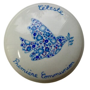 May include: White ceramic box with a blue and red floral patterned dove with a branch in its beak. The text "Celeste Premiere Communion" is written around the edge of the box.