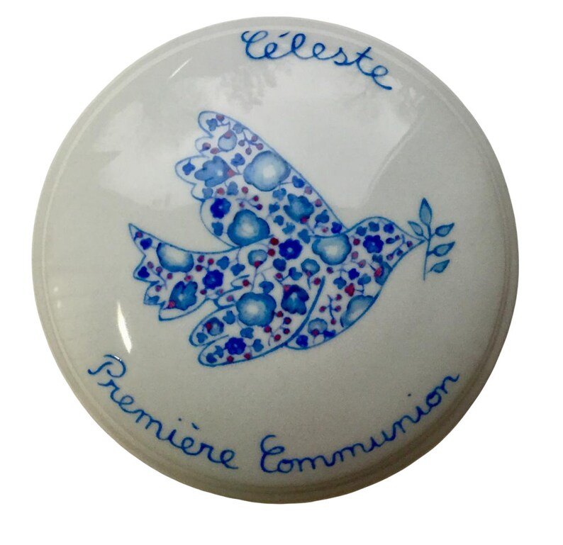 May include: White ceramic box with a blue and red floral patterned dove with a branch in its beak. The text "Celeste Premiere Communion" is written around the edge of the box.