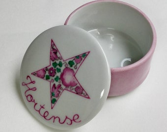 May include: A white porcelain trinket box with a pink lid. The lid features a pink and green floral star design and the name "Hortense" written in pink.