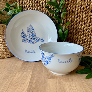 Peut inclure: Ensemble d'assiette et de bol en céramique blanche avec des motifs de colombe et de fleurs bleues. L'assiette présente une colombe portant un rameau d'olivier, et le bol a un motif similaire. Les deux pièces ont des bords bleus et le nom "Basile".