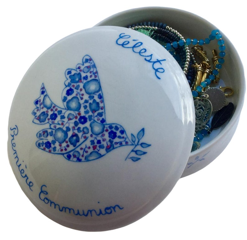 May include: White ceramic keepsake box with a blue and pink floral dove design. The lid has the words "Premi&egrave;re Communion" and "Celeste" written on it. The box is open and contains jewelry.