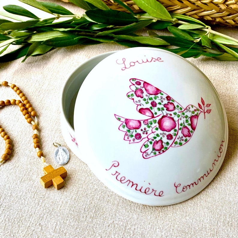 May include: A round white ceramic keepsake box with a lid featuring a hand-painted pink floral dove and the text Louise Premiere Communion. Beside the box lies a wooden bead rosary with a small silver medal and a wooden cross pendant.