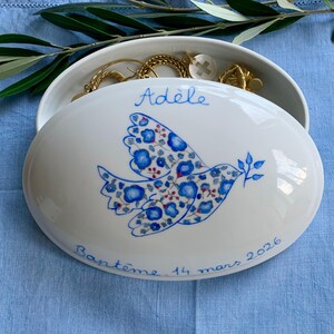 May include: White oval ceramic jewelry box with a blue floral dove design. The lid is inscribed with the name "Ad&egrave;le" and the date "14 mars 2026". The box is open, revealing gold jewelry inside.